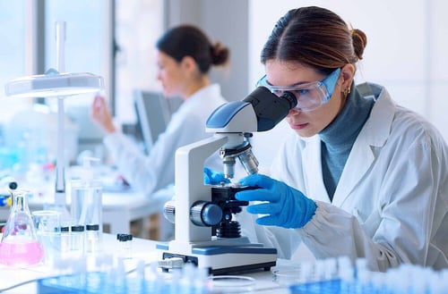 A woman in a lab coat looking into a microscope, working in a laboratory setting.