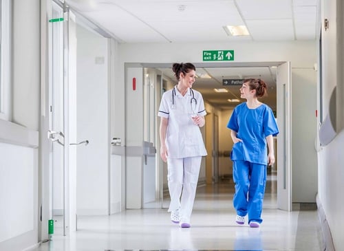 Two women walking down a hospital hallway, engaged in conversation, in a professional healthcare setting.
