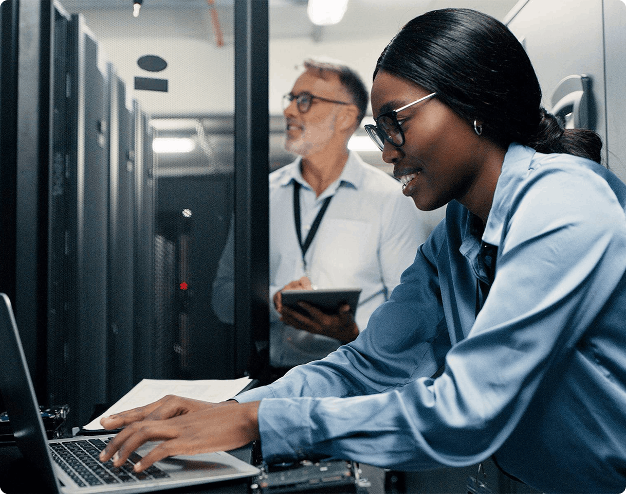 woman typing on laptop with man in background using tablet